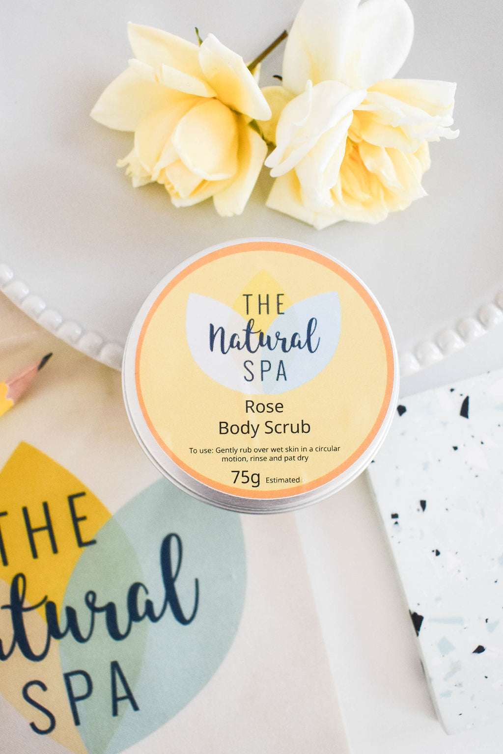 The Natural Spa Rose Sugar Scrub in stylish packaging on a decorative surface with yellow flowers.